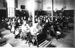 Class photo of students at Central School. The students are sitting in their desks. The teacher is at the back of the room. Note the music on the chalkboards.