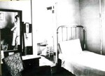 A bedroom inside the Young Women's Christian Association, YWCA, showing a single bed, dresser, bathroom, mirror, and stool. Note the woman's reflection in the mirror. The YWCA was located on Howell Avenue in Bisbee.