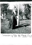 An actress in the play The Old Maids Convention. Depicts woman wearing a costume, entitled The Old Maids Convention. The play was also called the Spinsters Convention. She is holding a walking stick and smoking a pipe in a garden. She would have probably played the stereotypical spinster suffragette. The picture was taken in the Young Women's Christian Association, YWCA, garden. The Bank of Bisbee can be seen in the background. The YWCA was located on Howell Avenue in Bisbee.