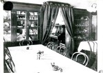 This photograph is identified as dining room 1908. This is the dining room of the old Young Women's Christian Association, YWCA, in Bisbee. There are two long tables with chairs. There is a sugar bowl, several salt and pepper shakers, a cruet and some flowers on the table. In the background is a large built-in china cabinet and a built-in cabinet holding a mineral collection. There is a curtained doorway between the two cabinets with a piano on the far side of the doorway. It also shows a wall lamp with glass globes.