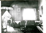 This photograph is identified as parlor or reception room - looking into office. 1908. It shows several rocking chairs, a piano, ceiling and wall lights, square table with a cloth on it, and a small desk with a straight chair. There is a fancy shade on the window. The office is seen through a curtained doorway, curtains are pulled open. The office seems to be the room seen in 1890.49.56. A desk with a woman sitting at it can be seen through the doorway. This is the interior of the old Young Women's Christian Association, YWCA, in Bisbee.
