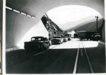 Photograph taken from inside the Mule Mountain Tunnel, looking west, showing the cars going through. It looks like only two of the three lanes are open. The photograph is dated 1957.