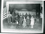 Photograph of the cast of a play standing on the stage at the Orpheum Theater in 1927. Back row, left to right: Marko Vucinich, Christ Papen, Chris Bugen, Sam Medigovich, George Thomasovich(?), Sam Vucotich, Marko Davidovich, Sam Sorich, and Lazai Martinovich. Front, left to right: R. Kasaticza(?), Gliso Chukovich, Ljubetca Cheranich, Petor Vuleksovich, and Mamie Bugen.