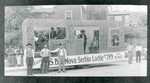 Photograph of the Serbian Lodge's float for the 1925 Fourth of July parade. The formal name of the lodge is on the float but is obscured in places by people. People are identified as: Archie Bugen, Sam Medigovich, Balich, Gliso Chukovich, Panto Malovich, Sizentich(?) in window, Masko Davidovich(?) sitting, and Mato Sklender. In window at left of picture: Chris Bugen, Steve Medigovich.