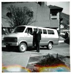 A 1970s Dodge Van used for transportation by the YWCA with driver Jay (?) Lirin(?) standing in front of the vehicle. The photo was taken in front of the YWCA building on Howell Avenue, with the Presbyterian Church and Copper Queen Hotel in the background. The wording on the side of the van is partially visible and reads Bisbee YWCA Services.